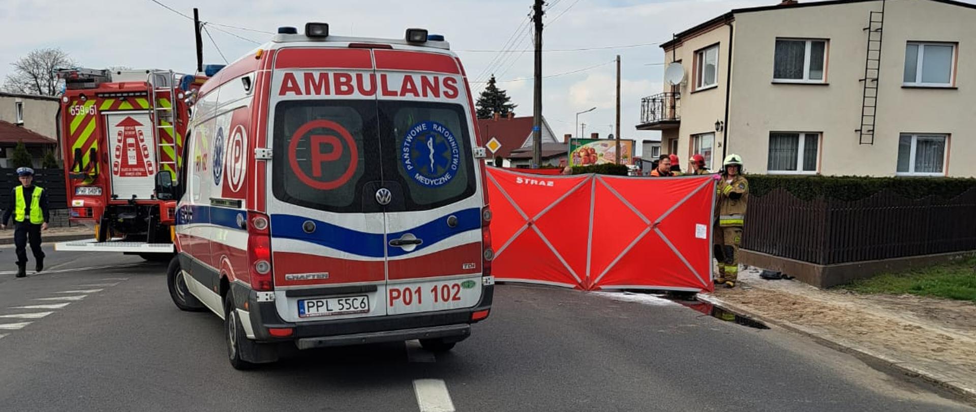 Zdjęcie przedstawia karetkę pogotowia, po jej prawej stronie czerwony parawan z napisem straż, a za nim stojących strażaków. Po lewej stronie wóz strażacki oraz policjant.
