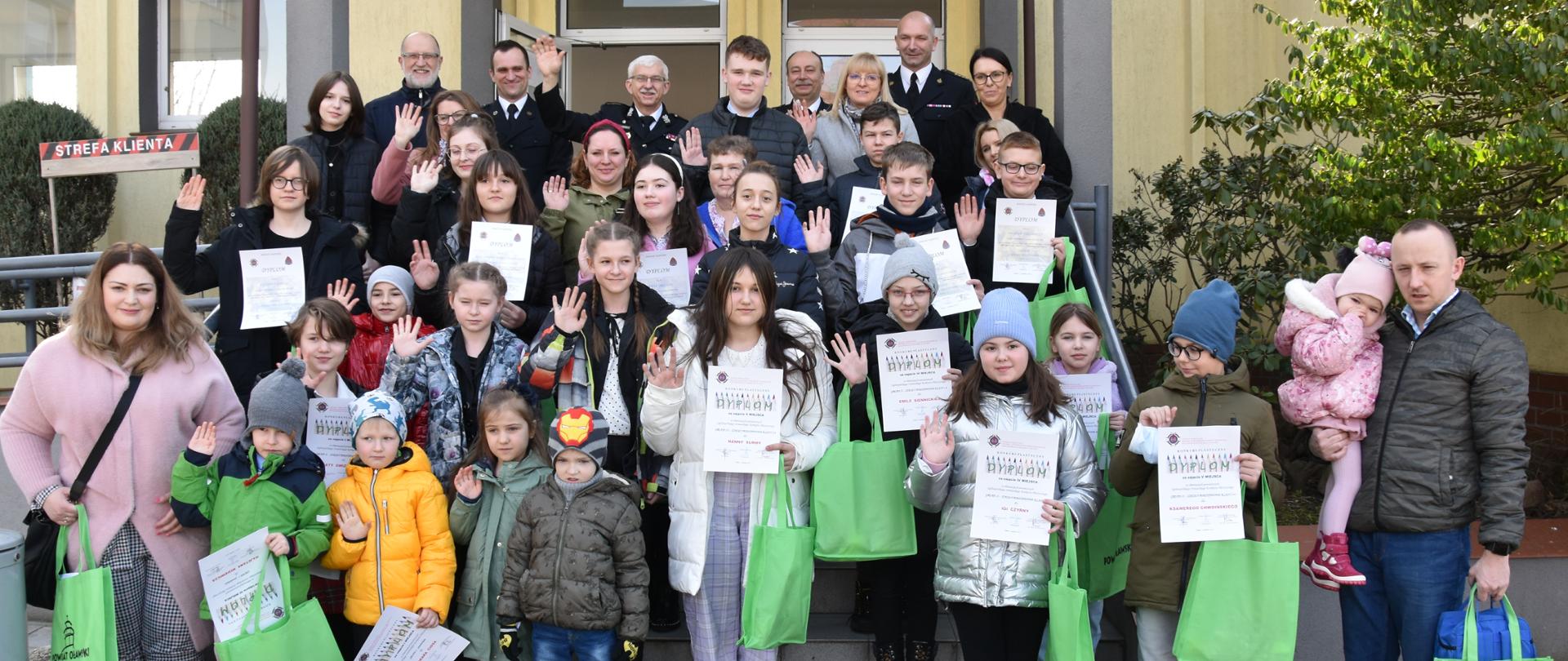 na zdjęciu widać grupę dzieci ze szkół podstawowych i średnich, z dyplomami i nagrodami. Dzieci zostały wyróżnione za udział w strażackich konkursach w ubiegłym roku. Wśród nich jest też starosta oławski, komendant powiatowy straży pożarnej oraz prezes ochotniczej straży pożarnej.