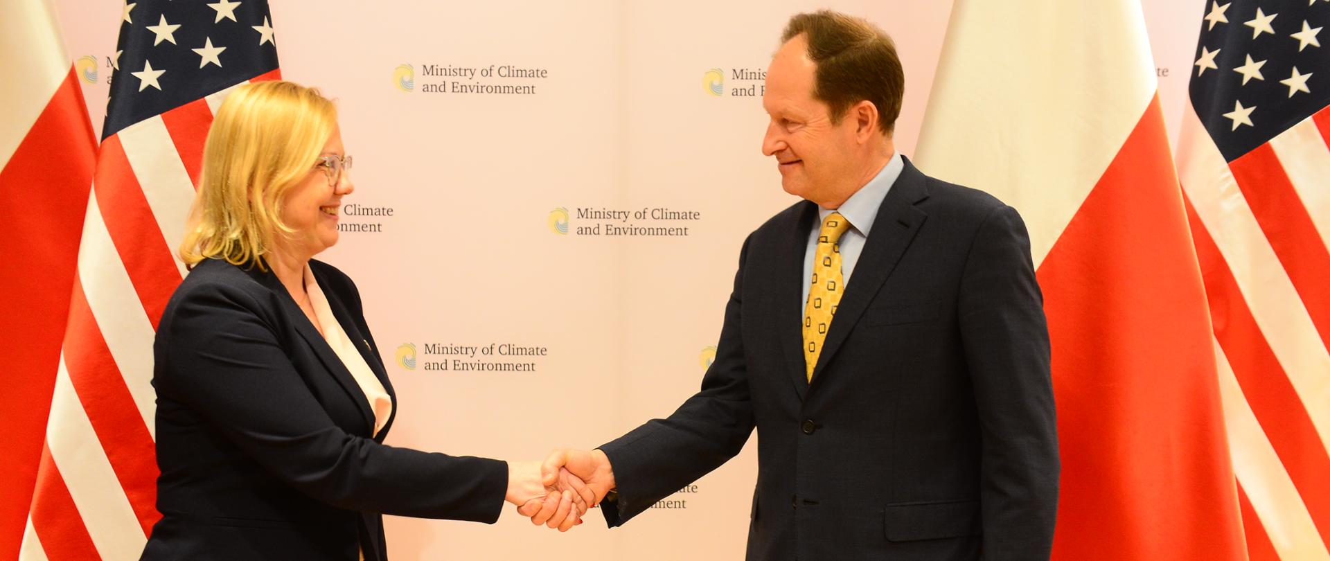 Minister Anna Moskwa ściskająca dłoń z Ambasadorem USA w Polsce Markiem Brzezinskim na tle flag Polski i USA