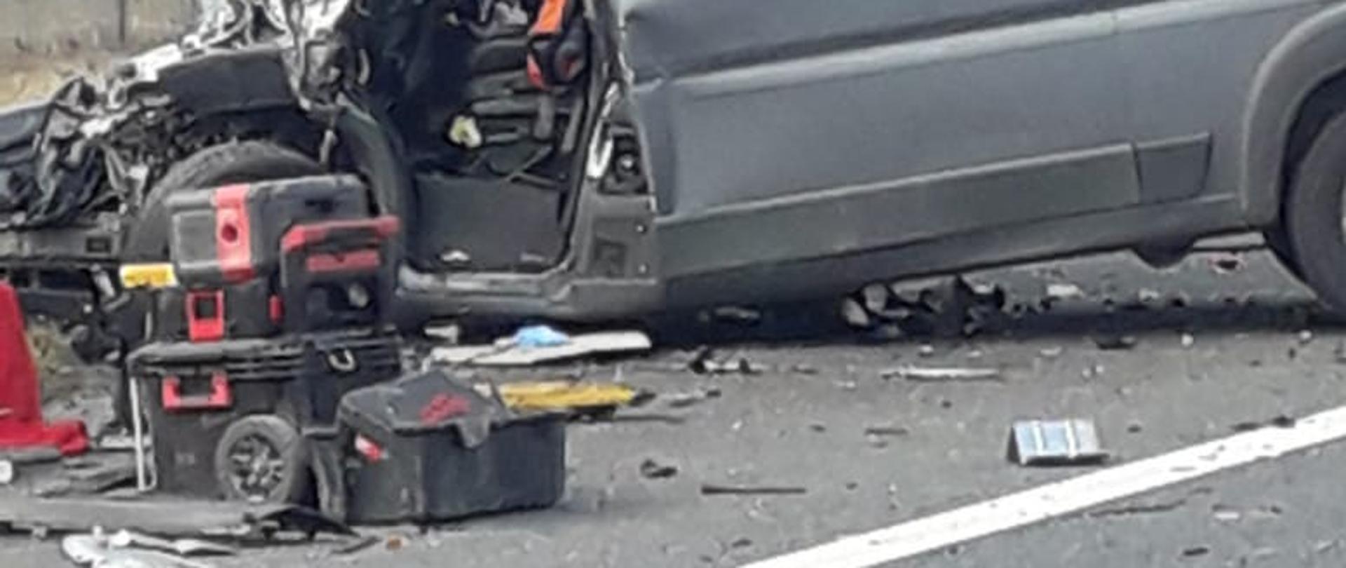 Na zdjęciu widoczny jest rozbity samochód dostawczy koloru szarego obok pojazdu złożone są skrzynki narzędziowe z tyłu pojazdu widoczny wiadukt drogowy. 