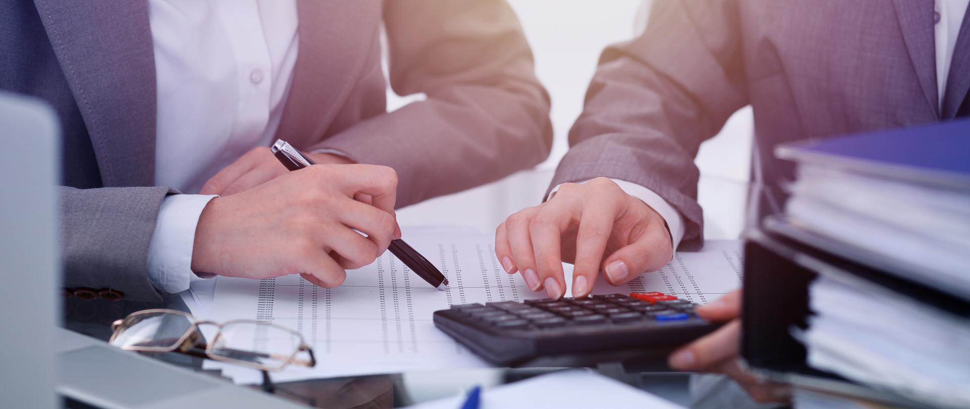 Dwie osoby przeliczają dane na kalkulatorze pomiędzy laptopem a segregatorami
