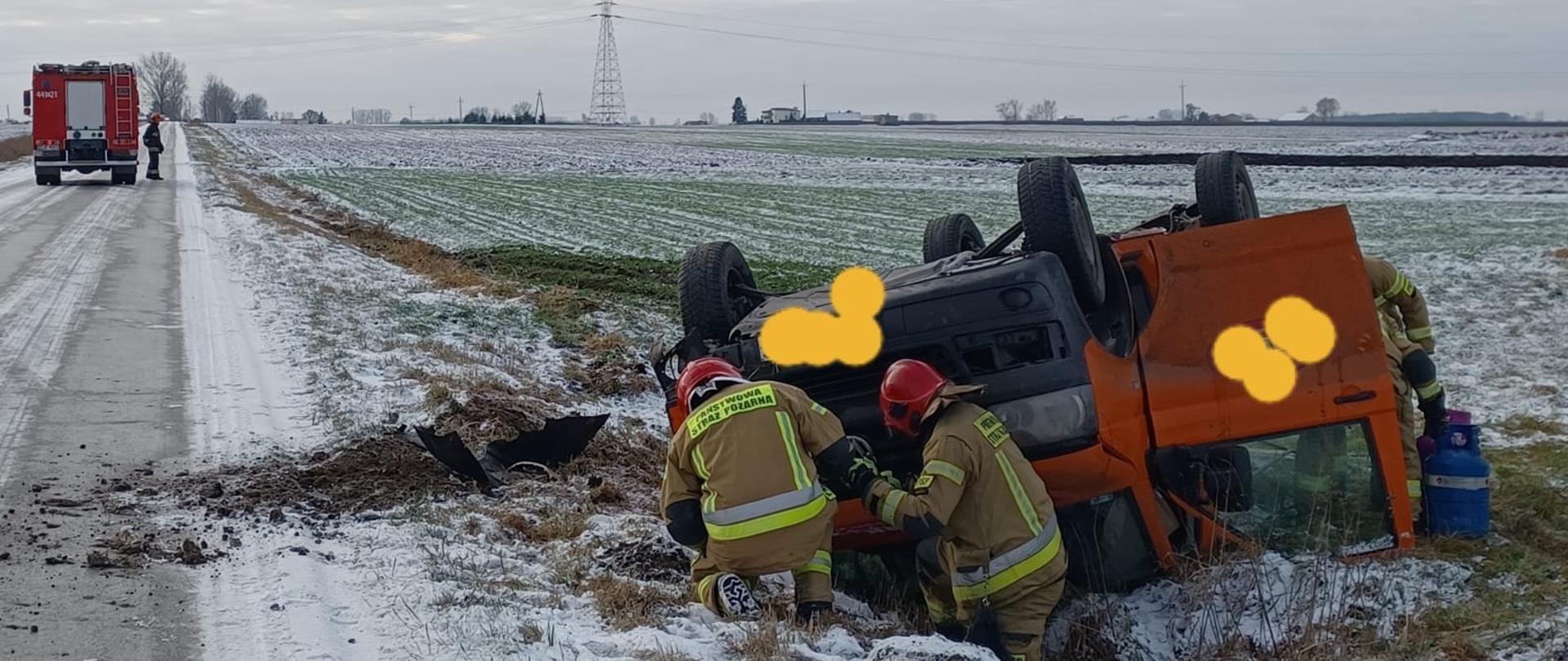 Na zdjęciu widać pomarańczowy samochód dostawczy, który leży w rowie kołami w górze. Obok samochodu widać czterech strażaków w żółtych strojach i czerwonych hełmach. Obok stoją dwie butle z gazem. W tle widać na drodze czerwony samochód strażacki, zabudowania, pola, krzewy i drzewa. Na ziemi jest cienka warstwa śniegu. Jest widno.
