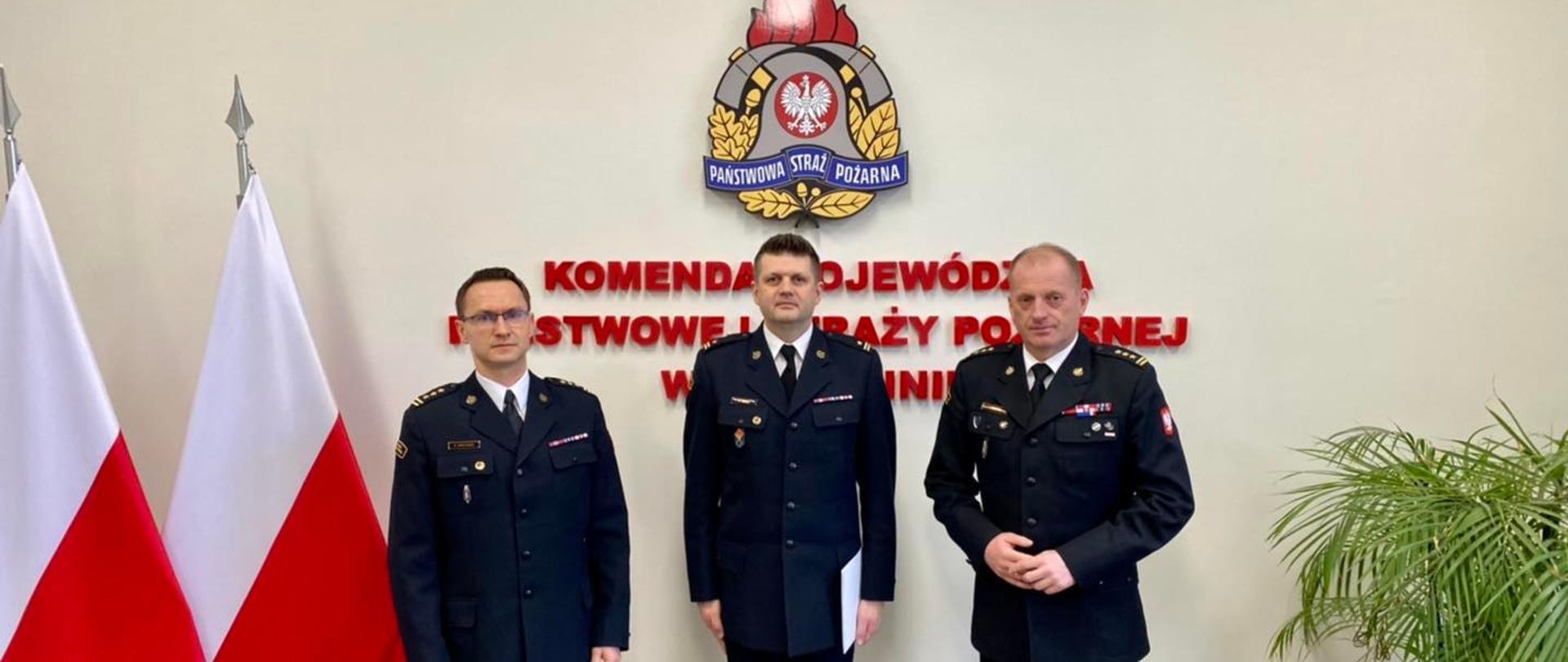 Na zdjęciu znajduje się trójka mężczyzn, po lewej stronie flag. Po prawej stronie zielona roślinka.
W tle logo PSP wraz z napisem.