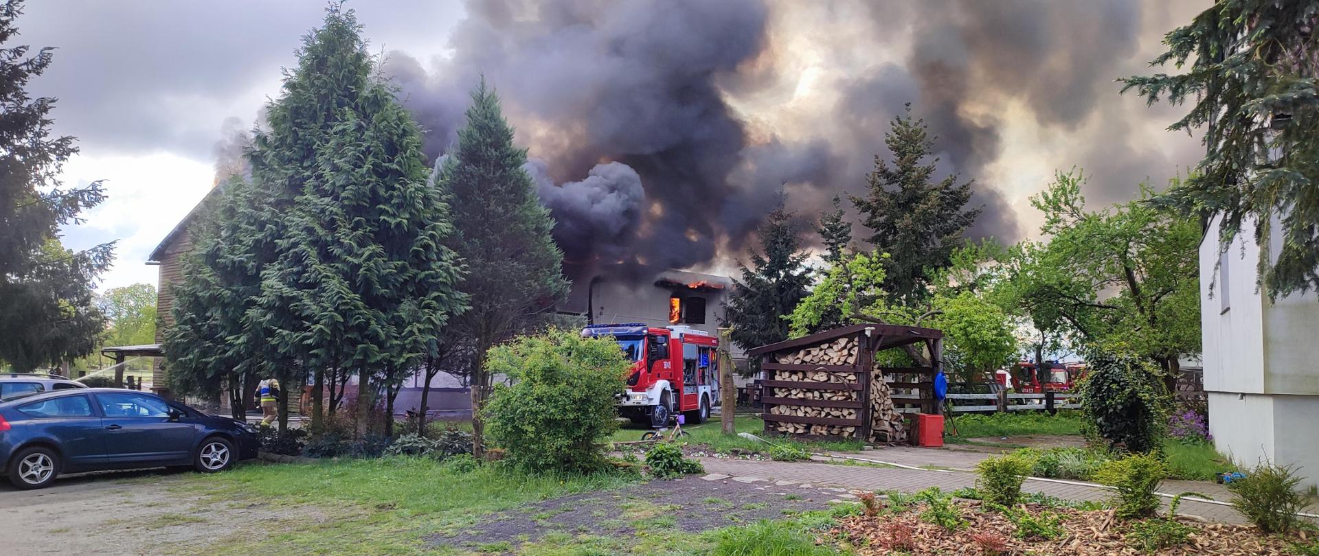 Zdjęcie przedstawia objęty pożarem budynek stajni, z którego wydobywają się kłęby czarnego dymu. W tle znajdują się pojazdy pożarnicze.