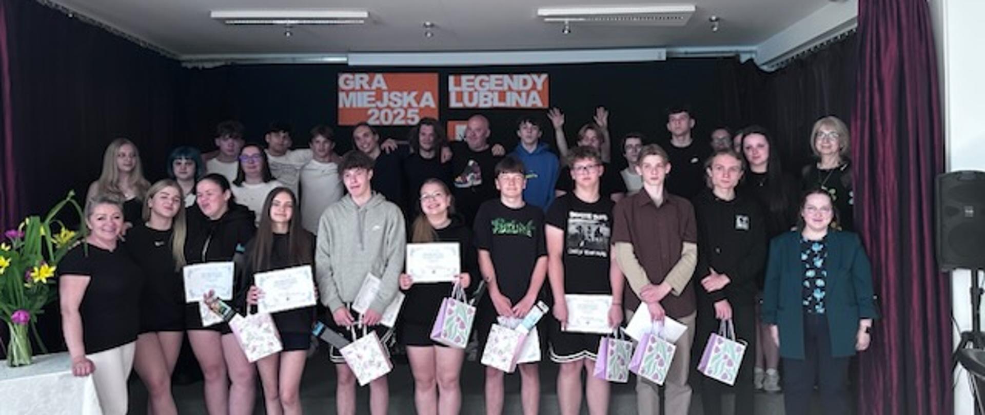 W pomieszczeniu na czarnym tle stoi przodem grupa kilkudziesięciu osób. Są uśmiechnięci, osoby z przodu trzymają białe kartki i papierowe torby biało-różowe. Za nimi na górze znajduje się biały napisz na pomarańczowym tle : ""GRA MIEJSKA 2025 LEGENDY LUBLINA".