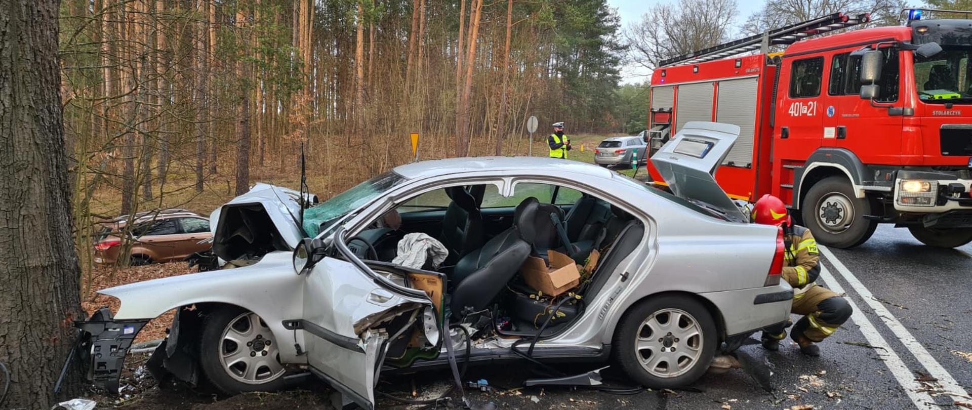 Zdjęcie przedstawia widok lewego boku rozbitego srebrnego samochodu osobowego marki Volvo, nadwozie sedan. Samochód z całkowicie zgniecionym przodem stoi przy drzewie rosnącym na poboczu. Tył pojazdu stoi na jezdni. W samochodzie jest wycięty środkowy słupek wraz z tylnymi drzwiami, drzwi kierowcy są otwarte, częściowo pogięte. Wewnątrz pojazdu widać uruchomione poduszki powietrzne kierowcy i pasażera, fotel kierowcy jest w pozycji leżącej. Dalej za rozbitym Volvo na całej szerokości jezdni stoi samochód gaśniczy strażacki, blokując ruch. W tle widać las porastający pobocze, inne pojazdy osobowe oraz policjanta. Przy otwartej tylnej klapie Volvo kuca strażak ubrany w ubranie specjalne i hełm, robi coś wewnątrz bagażnika. Ujęcie wzdłuż drogi krajowej numer 22. Jest dzień, widno.