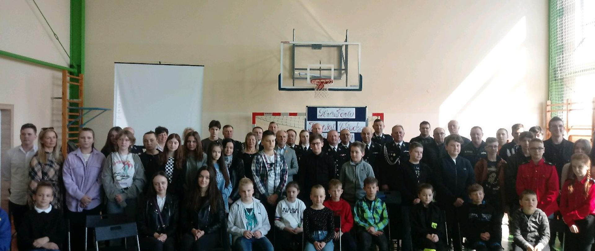 Na zdjęciu widać umundurowane osoby w pomieszczeniu. Strażacy OSP i PSP oraz osoby cywilne, dzieci. Pomieszczenie szkolne. Zdjęcie wykonane w ciągu dnia.