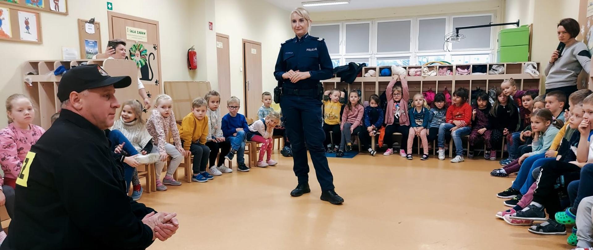 Na zdjęciu znajdują się dzieci oraz strażak ubrany w ubiór służbowy i policjantka, w sali podczas pogadanki