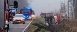 Na zdjęciu widać miejsce wypadku drogowego. Na pierwszym planie, po lewej stronie, stoi częściowo widoczny czerwony wóz strażacki. Obok niego, na pasie ruchu, zatrzymał się srebrny samochód osobowy marki Skoda. Za nim stoi kilka innych pojazdów, w tym co najmniej dwa czerwone wozy strażackie z włączonymi niebieskimi światłami ostrzegawczymi.
Po prawej stronie drogi, w przydrożnym rowie, leży przewrócony na bok ciągnik siodłowy z białą cysterną. Wokół przewróconego pojazdu stoi grupa strażaków w żółtych ubraniach interwencyjnych i czerwonych hełmach. Nad miejscem wypadku unosi się mgła, a w tle widać drzewa.