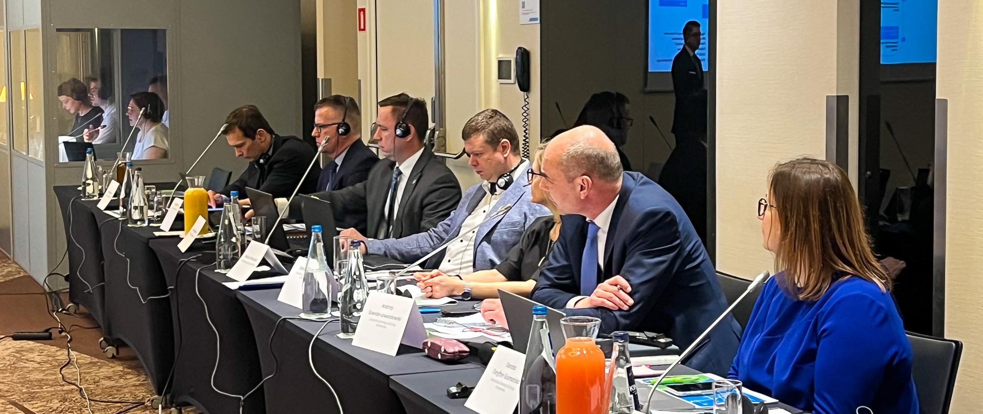 Sala konferencyjna. Uczestnicy konferencji (kobiety i mężczyźni) siedzą za stołem. Przed nimi laptopy i dokumentacja. 