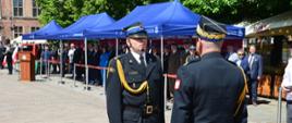 Strażak w mundurze galowym melduje strażakowi w mundurze galowym obok pod niebieskimi namiotami stoją zaproszeni goście oraz przedstawiciele służb mundurowych obok znajduje się mównica.