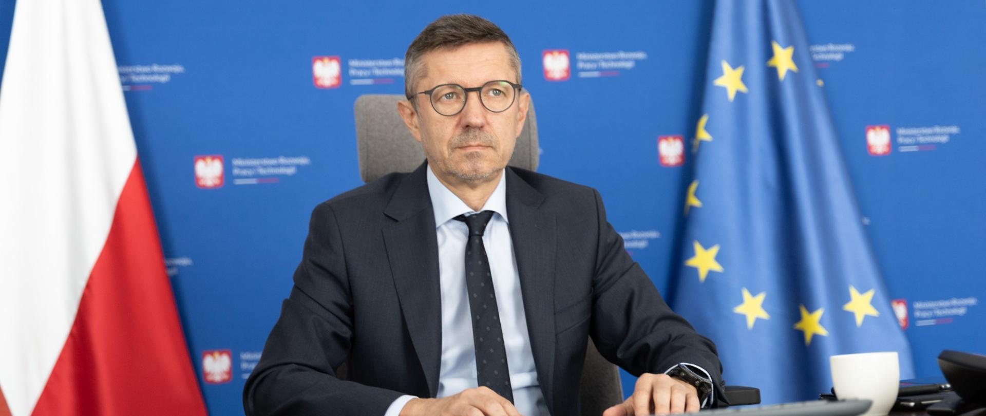 Wiceminister Robert Tomanek na tle flag Polski i UE oraz ścianki z logotypem MRPiT.