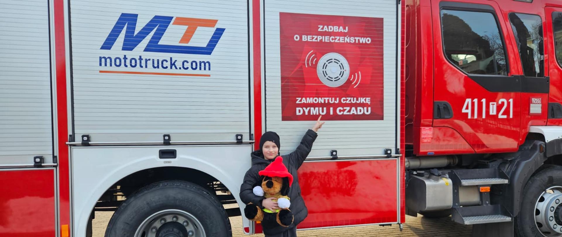Na zdjęciu chłopiec, lat 10 trzymający w rękach Misia Ratownika i palcem wskazuje na plakat „Zadbaj o bezpieczeństwom Zamontuj Czujkę Dymu i Czadu” na samochodzie ratowniczo-gaśniczym