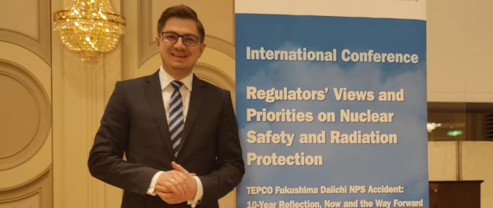 Prezes PAA na konferencji dot. priorytetów w bezpieczeństwie jądrowym - dr Łukasz Młynarkiewicz stoi obok baneru promującego konferencję. 