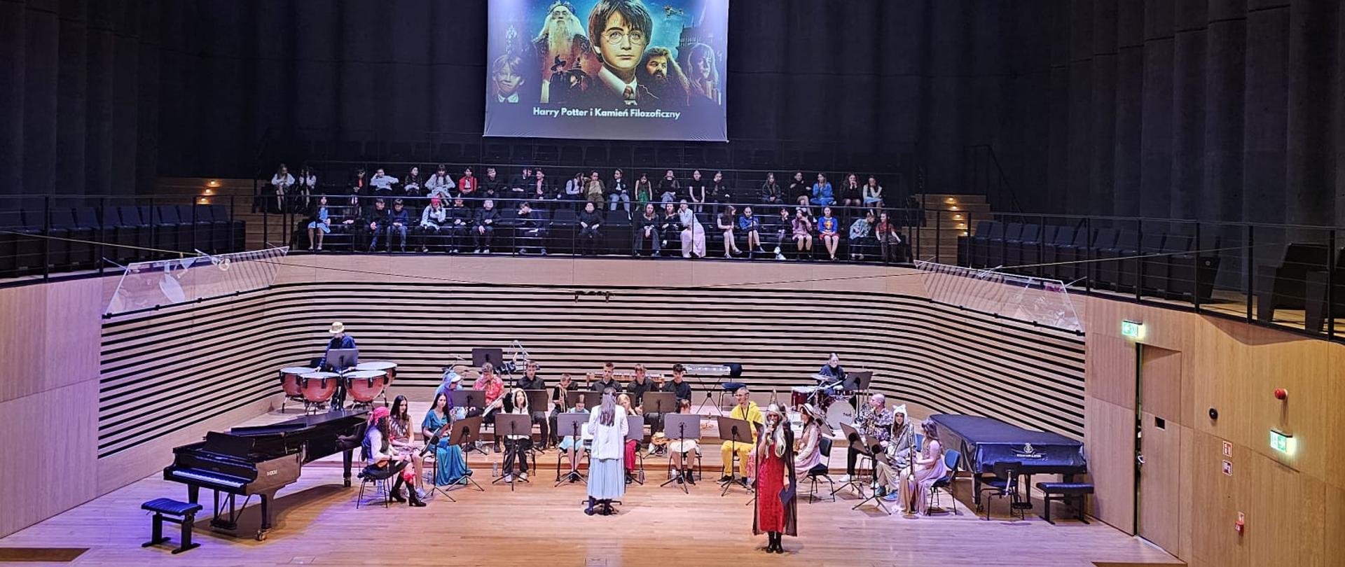 Kobieta w przebraniu Czarodziejki mówi do mikrofonu, za nią siedzi orkiestra i stoi kobieta odwrócona tyłem do publiczności.