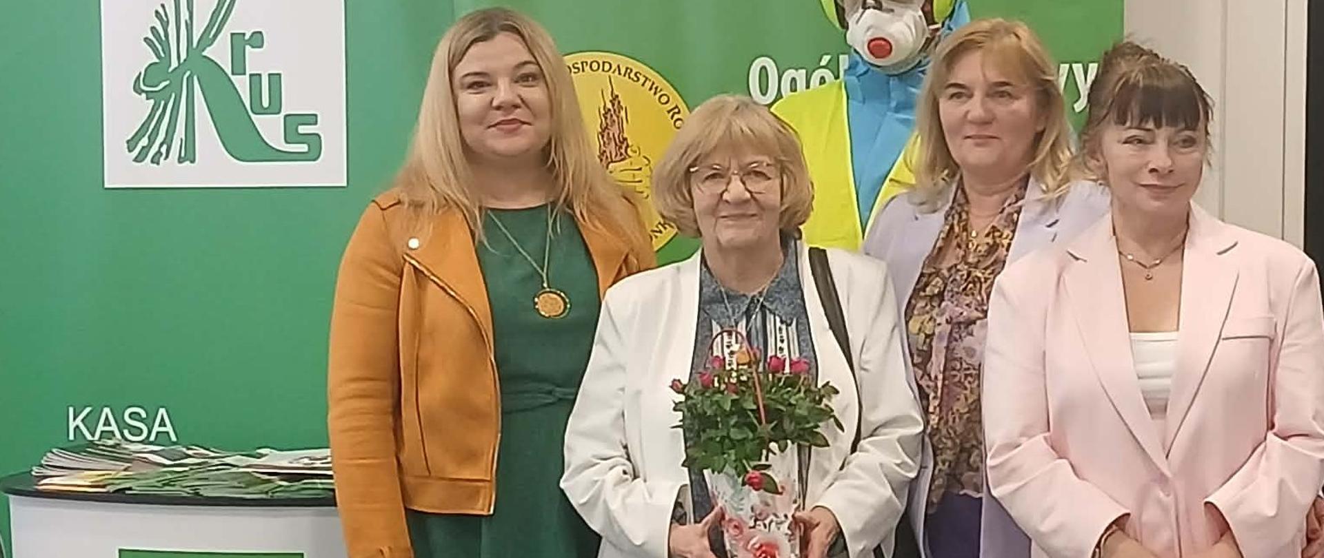 Cztery kobiety pozują do zdjęcia stojąc na tle zielonej ścianki z logo KRUS, przy pulpicie reklamowym KRUS. Dwie z nich trzymają w rękach kwiatki doniczkowe w ozdobnych torebkach.