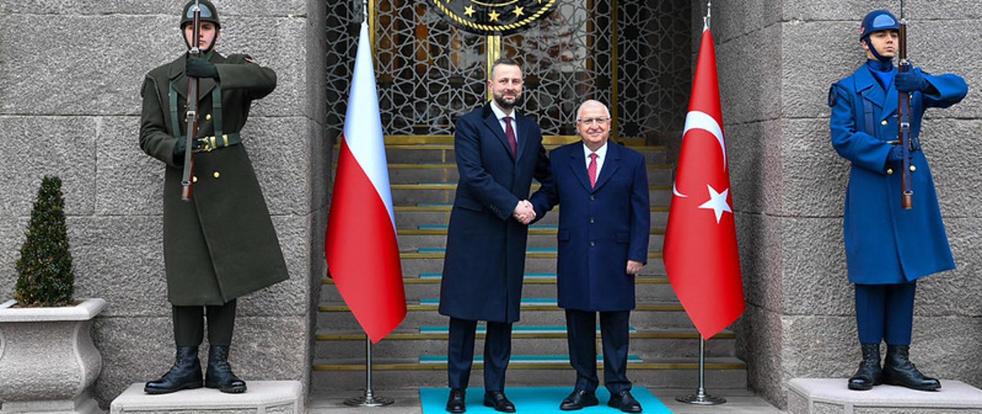 Dwóch mężczyzn stoi na schodach przed oficjalnym budynkiem i podaje sobie dlonie. Po obu stronach widoczne sa flagi Polski i Turcji.