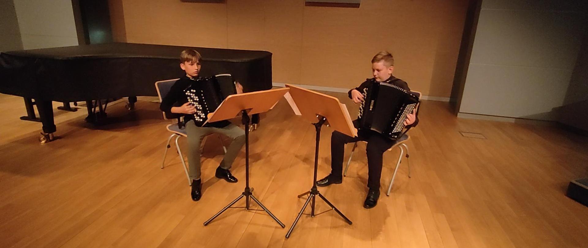 Zdjęcie uczniów siedzących na krzesłach, z instrumentami, przy pulpitach. W tle fortepian oraz prospekt organowy. Zdjęcie wykonane na scenie Sali Koncertowej ZPSM im. F. Chopina w Jarosławiu