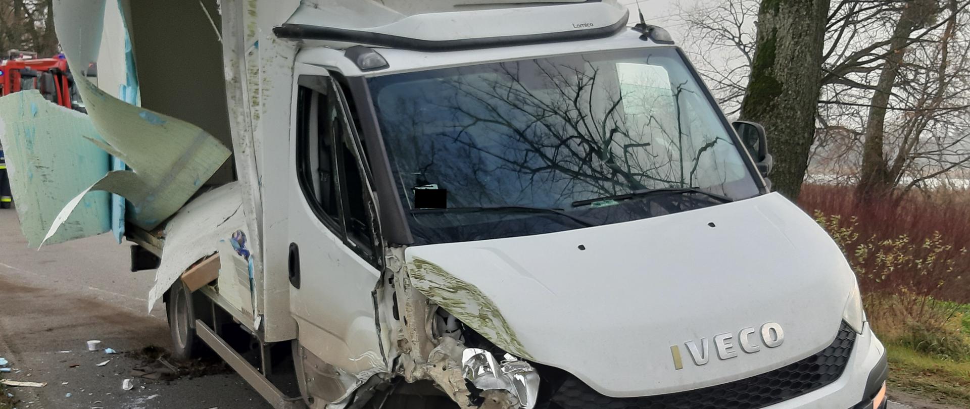 Biały samochód dostawczy stoi na drodze. Ma rozbity przód i uszkodzony bok zabudowy, za nim stoi samochód gaśniczy. 