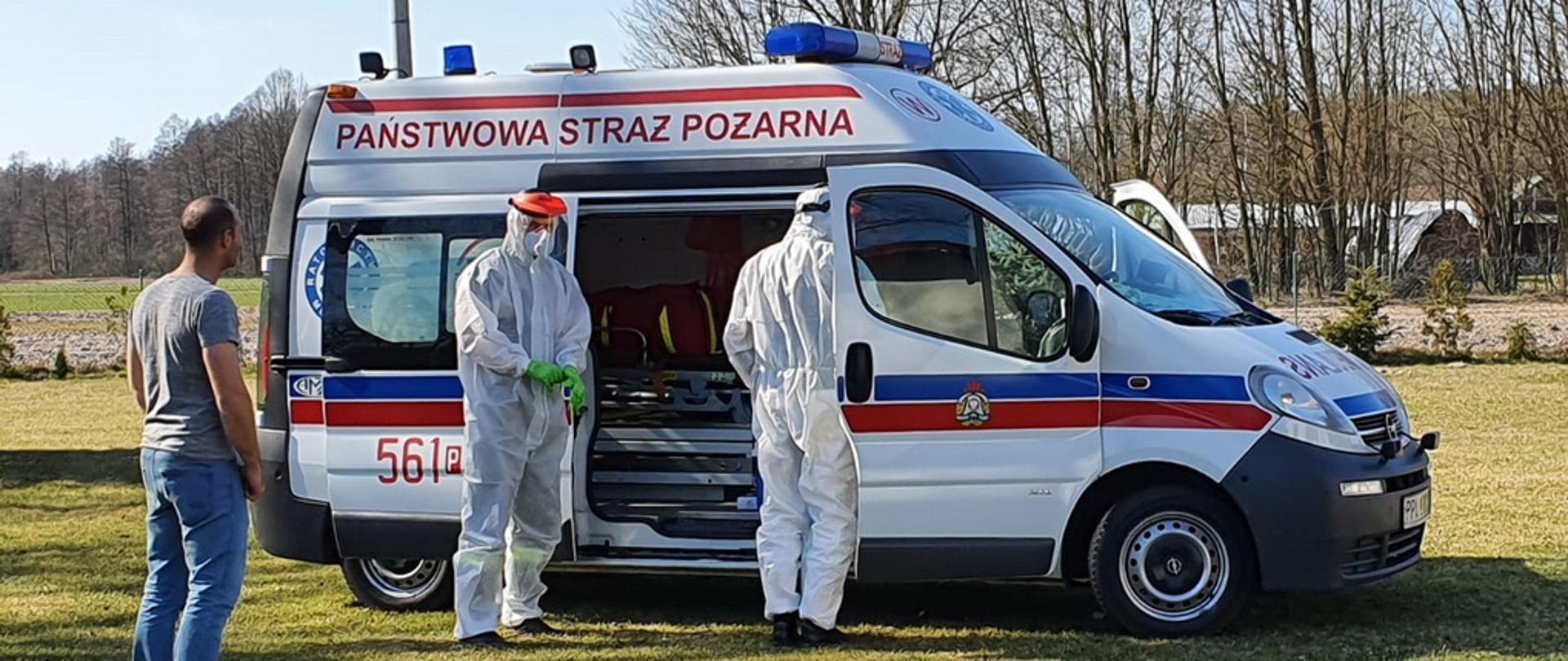Pobór próbek strażacy na tle strażackiej karetki z KP PSP Pleszew i stojąca w bezpiecznej odległości osoba po pobraniu próbki