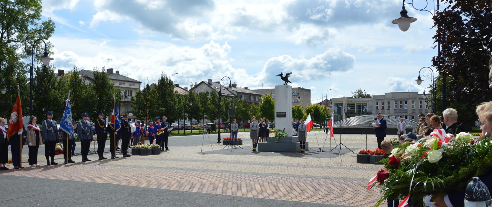 W parku miejskim pod pomnikiem Ofiar II wojny światowej zgromadzeni są ludzie. Po lewej stronie stoją poczty sztandarowe wraz z orkiestrą . Po prawej stronie stoją zaproszeni goście z wiązankami kwiatów oraz zniczami. W centralnej części zdjęcia pod pomnikiem stoją dwie kolumny z głośnikami, harcerze oraz mężczyzna przemawiający przez mikrofon.
