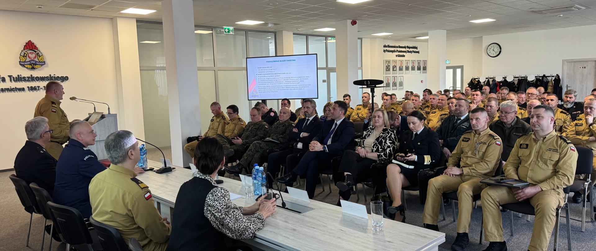 Uczestnicy narady rocznej kadry kierowniczej Państwowej Straży Pożarnej województwa pomorskiego podczas obrad w sali konferencyjnej. Strażacy w umundurowaniu oraz zaproszeni goście siedzą naprzeciw stołu prezydialnego i słuchają wystąpienia prowadzącego spotkanie.