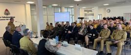 Uczestnicy narady rocznej kadry kierowniczej Państwowej Straży Pożarnej województwa pomorskiego podczas obrad w sali konferencyjnej. Strażacy w umundurowaniu oraz zaproszeni goście siedzą naprzeciw stołu prezydialnego i słuchają wystąpienia prowadzącego spotkanie.