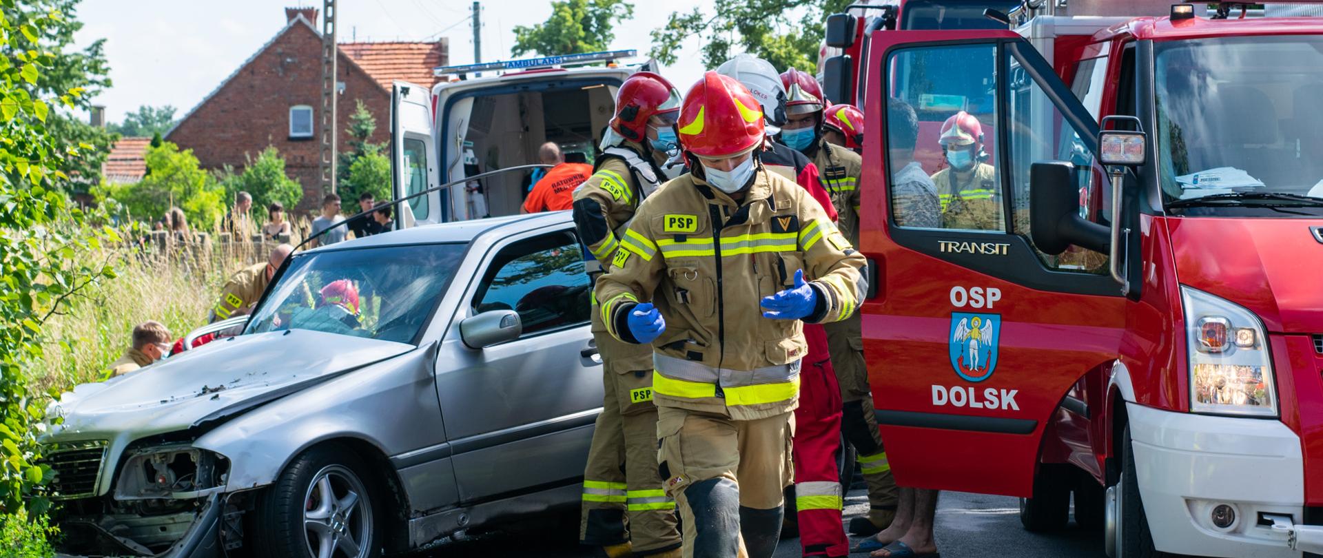 Na zdjęciu rozbity samochód osobowy, zespół ratownictwa medycznego oraz zastęp OSP Dolsk, strażacy i ratownicy medyczni podczas swoich czynności oraz strażak idący 