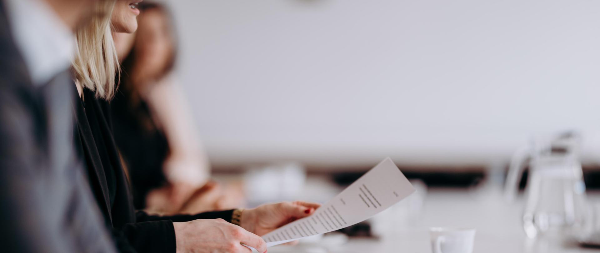 Spotkanie biznesowe, przy stole siedzą uczestnicy, ich twarze są niewidoczne. Na drugim planie kobieta trzymająca w ręce dokument. Na blacie stoją filiżanki i dzbanek
