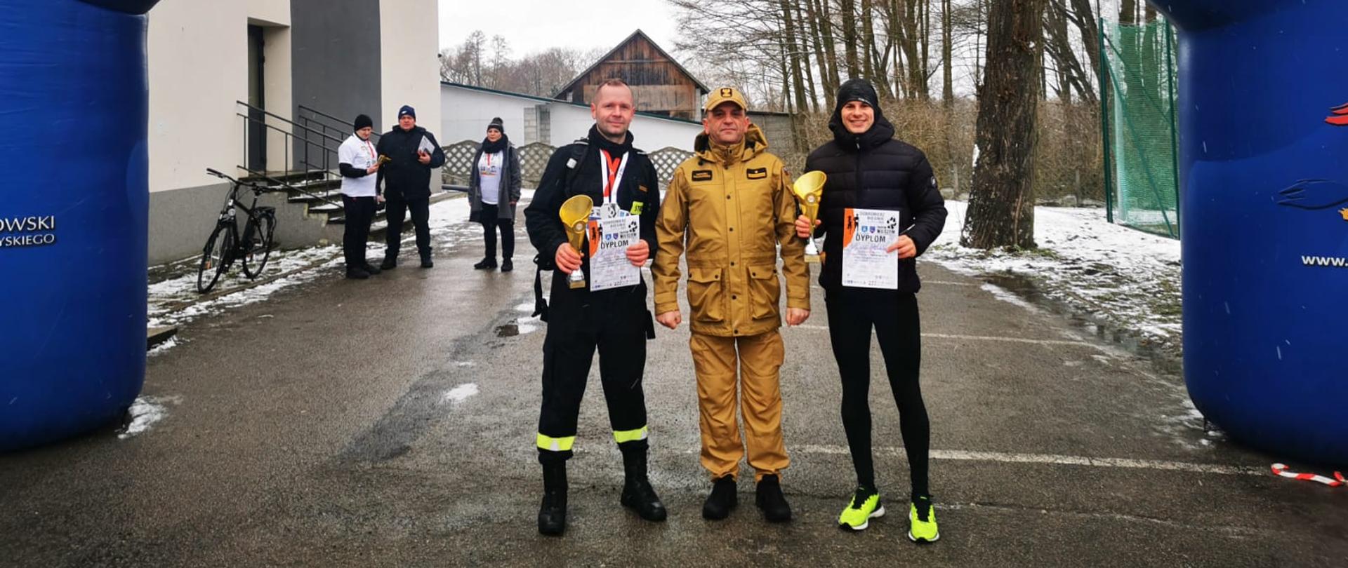 Teren szkoły podstawowej w Dobromierzu, pod pneumatyczną bramą startową stoją funkcjonariusze JRG Włoszczowa, w rękach trzymają trofea, pomiędzy nimi w umundurowaniu służbowym komendant powiatowy PSP we Włoszczowie.