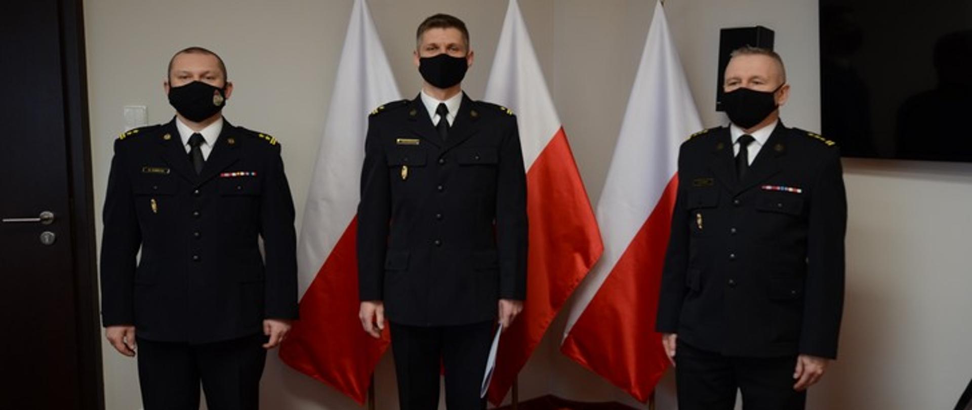 Zdjęcie przedstawia trzech oficerów PSP w mundurach wyjściowych na tle flag państwowych.