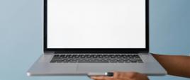 Dłonie trzymają otworzony laptop na niebieskim tle. Obudowa laptopa szaro-czarna Tło ekranu białe.