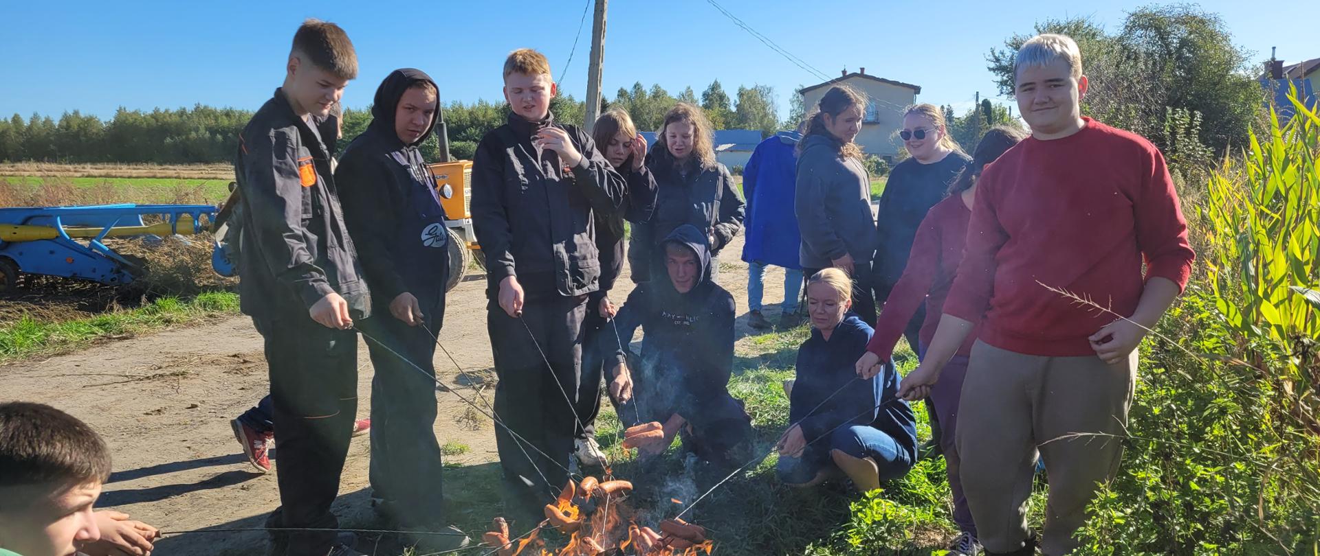 Grupa uczniów i nauczycielka wokół ogniska, smażą kiełbaski