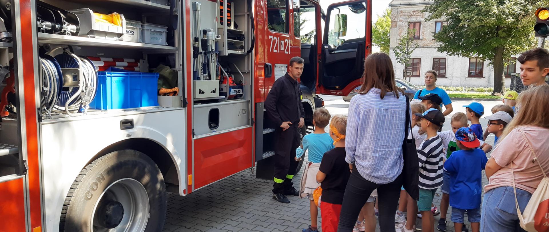 Grupa dzieci wraz z opiekunami stoi na dworze przy samochodzie strażackim. Strażak pokazuje im wyposażenie samochodu.