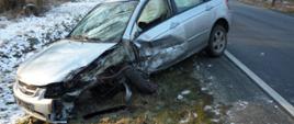 Na zaśnieżonym rowie stoi samochód osobowy koloru srebrnego z uszkodzonym przodem i bokiem, widać urwane koło i wystrzelone poduszki powietrzne. 