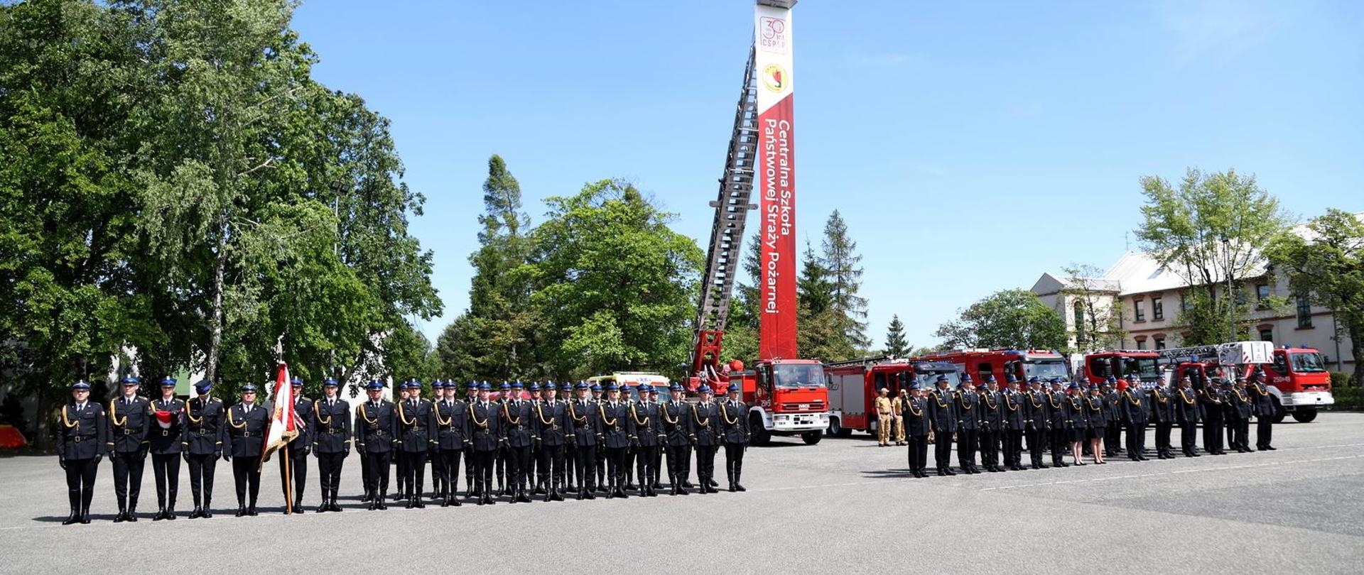 Pododdziały strażaków stoją na placu wewnętrznym Szkoły podczas uroczystości, w głębi samochód bojowy typy drabina z banerem zwisającym z drabiny