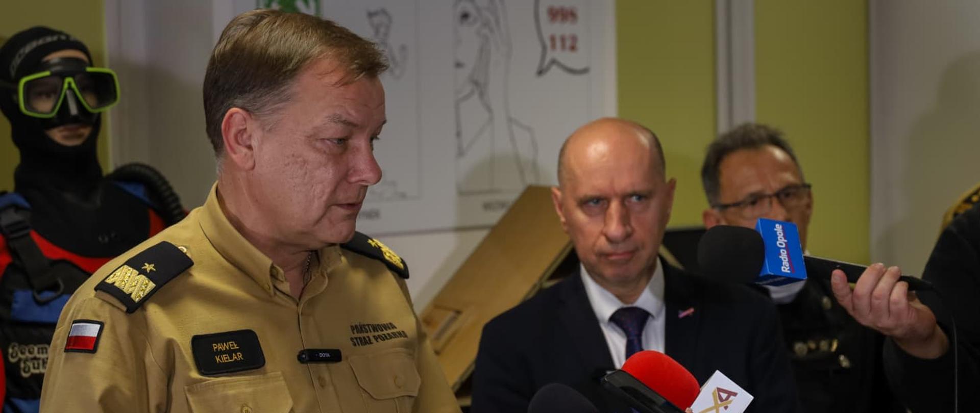 Nadbrygadier Paweł Kielar z Opolski Komendant Wojewódzki Państwowej Straży Pożarnej udziela wypowiedzi mediom podczas briefingu o zagrożeniach sezonu grzewczego. W tle manekiny ratowników i plakaty edukacyjne z numerami alarmowymi. Obok Wicewojewoda Opolski.