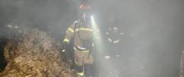 Dwóch strażaków wychodzi z zadymionego pomieszczenia obok nich widać słomę oraz elementy wyposażenia kurnika strażacy ubrani w aparaty ochrony dróg oddechowych z zapaloną latarka jeden trzyma w reku kamerę termowizyjną
