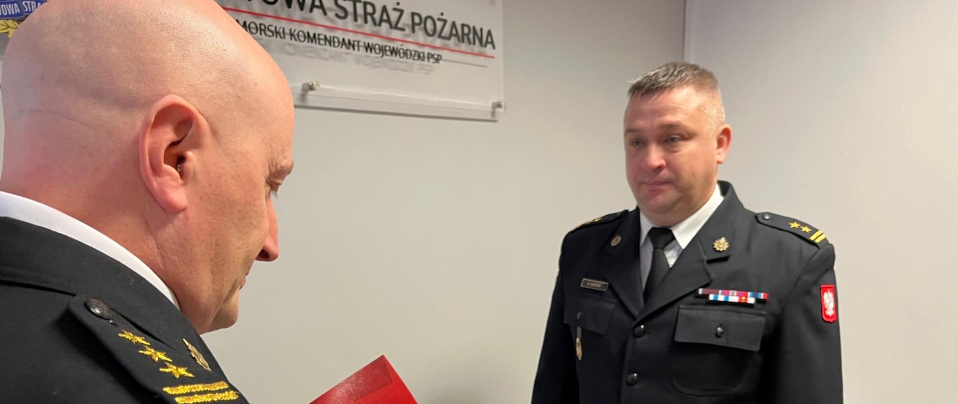 Dwóch funkcjonariuszy w mundurach wyjściowych stoi zwróconych do siebie twarzami. Ten z lewej odczytuje dokument. W tle po prawo brązowa komoda.