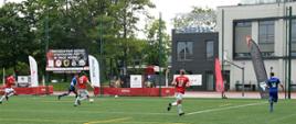 Zawodnicy w strojach niebieskich oraz biało-czerwonych grają w piłkę nożna na boisku niedaleko znajduje się budynek.