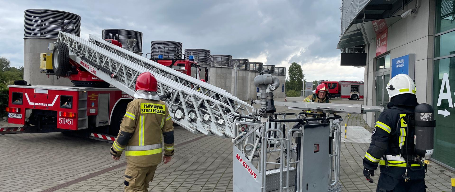 Na zdjęciu rozłożona drabina mechaniczna. Strażacy PSP i OSP z aparatami powietrznymi na plecach uzbrajają kosz drabiny w nosze i działko gaśnicze