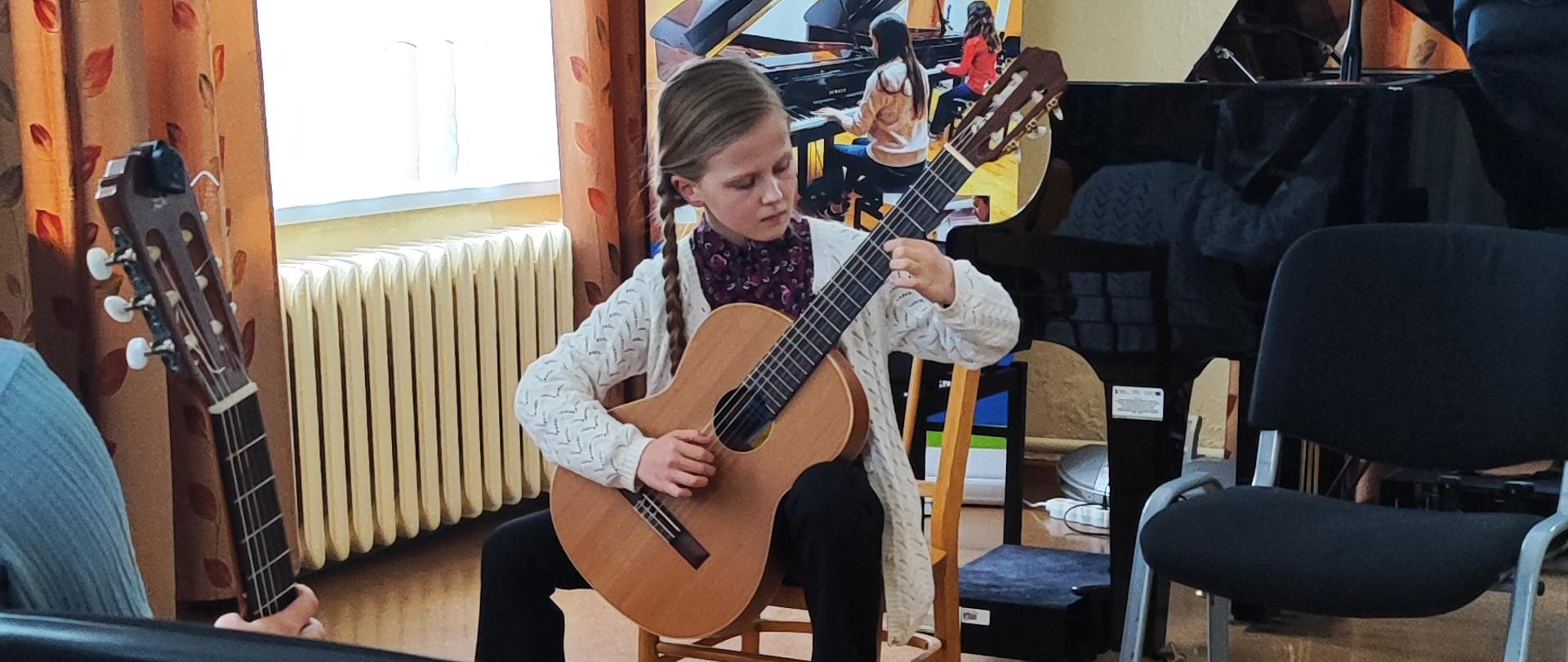 Uczennica na siedząco gra na gitarze za nim baner unijny, fortepian, ława fortepianowa, okno zasłonięte firaną i dwoma zasłonami, nad nim wisi lampa. Obok niej po prawej stronie puste krzesło. Pa pierwszym planie puste krzesło