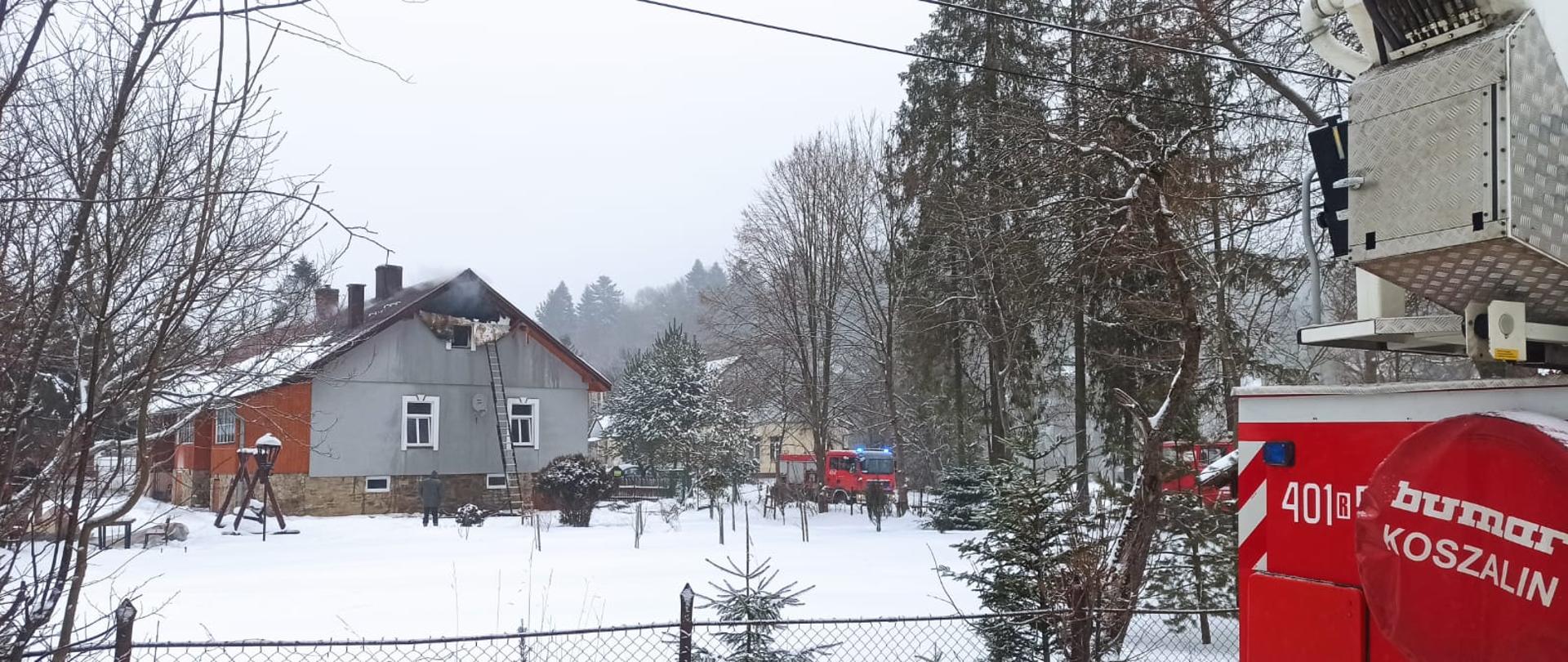 Na zdjęciu widać dom jednorodzinny z którego u szczytu dachu oderwana jest elewacja i wydobywa się dym. W miejscu tym jest też rozstawiona drabina 10 metrowa wysuwna. Zdjęcie zrobione jest przed ogrodzeniem działki po prawej stronie widać fragment samochodu SHD-25 oraz w oddali samochody Man TGM oraz Scania P400 Wszystkie te pojazdy należą do JRG w Ustrzykach Dolnych. Na działce rosną drzewa, przy domu stoi huśtawka oraz stół i ławki. Działka pokryta jest śniegiem. 