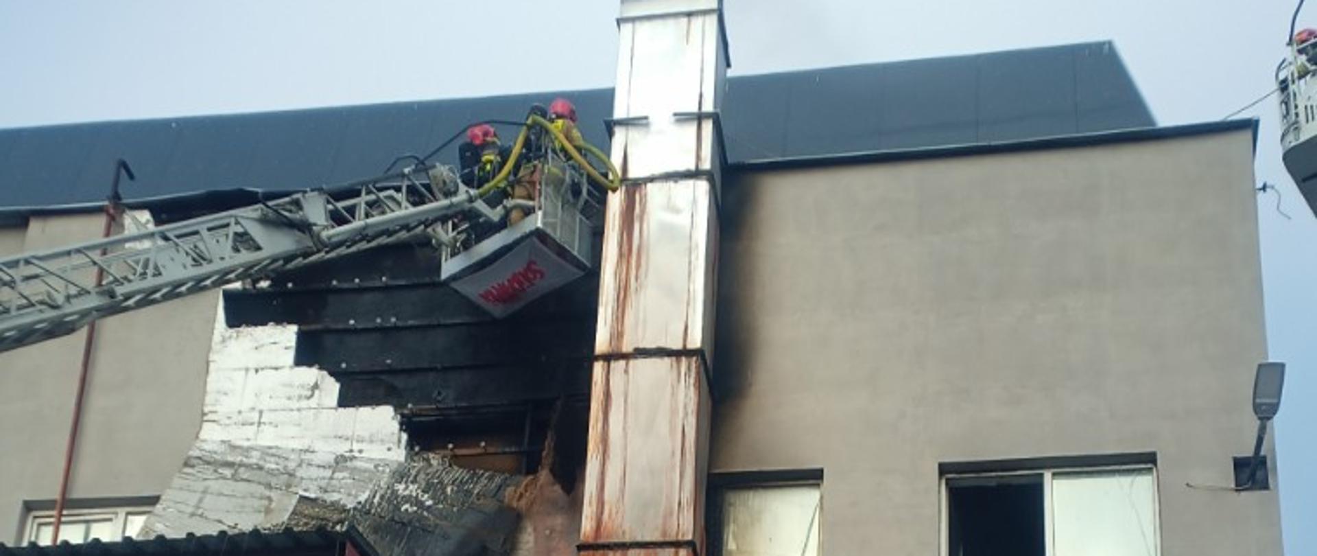 Strażacy w koszu drabiny mechanicznej na tle budynku z częściową spaloną elewacją 