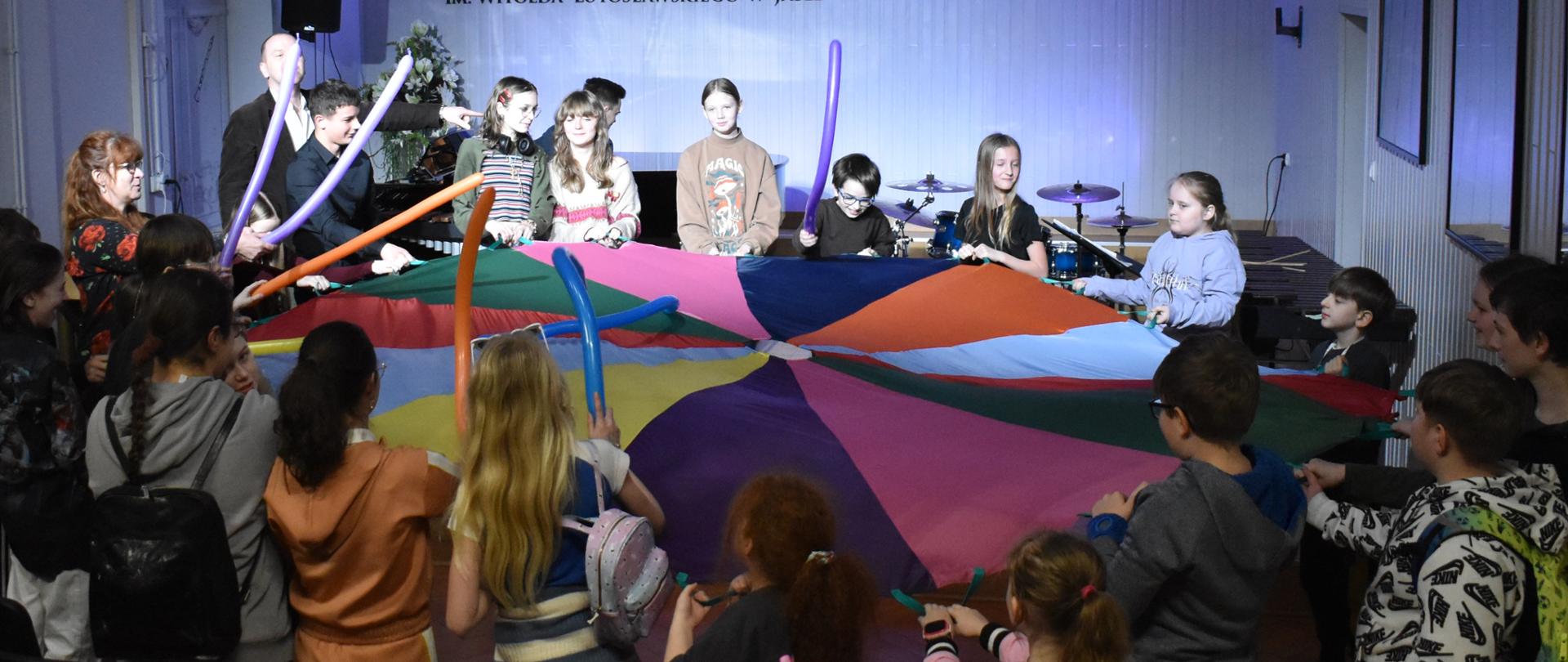 na zdjęciu, w auli PSM widnieje społeczność szkolna, rozciągająca kolorową płachtę, która ma obrazować największy bęben świata. Niektórzy uczniowie i nauczyciele uderzają w płachtę długimi, kolorowymi balonikami Kolorystyka zdjęcia jest biało-szaro-żółto-różowo-czerwono-zielono-pomarańczowo-brązowo-niebiesko-czarna.