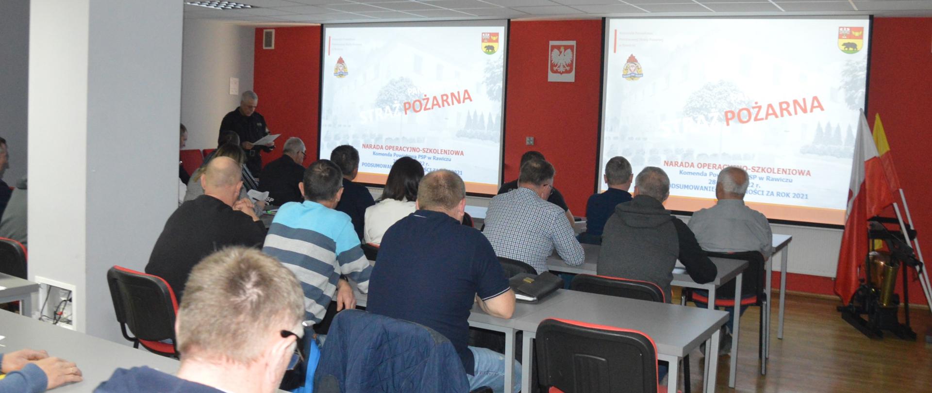 Narada operacyjno-szkoleniowa. W świetlicy komendy odbywa się narada z udziałem pracowników urzędów, komendantów gminnych i przedstawicieli OSP. Uczestnicy oglądają na ekranach przygotowaną prezentację multimedialną. naradzie przewodniczy zastępca komendanta powiatowego PSP. Z prawej strony znajduje się stojak z flagami.
