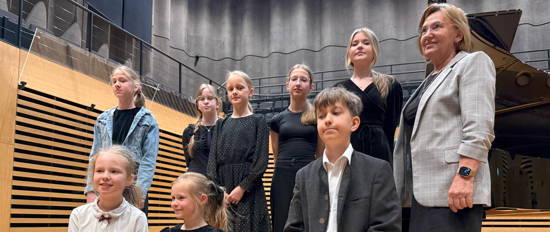 Uczniowie i kobieta stoją na scenie przy fortepianie.