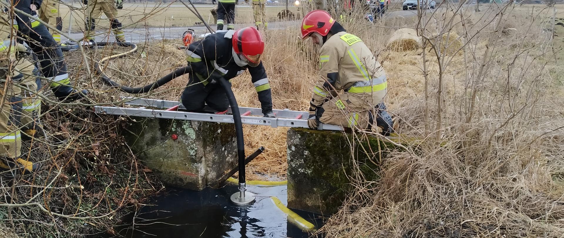 Fotografia przedstawia żelbetowy jaz pozbawiony przegrody na kanale melioracyjnym. Kanał szerokości około półtora metra. Brzegi kanału porośnięte gęstą suchą trawą. Kanał jest wypełniony wodą. Dwóch strażaków w ubraniach specjalnych i w hełmach klęczy na drabinie nasadkowej ułożonej na dwóch brzegach jazu. Strażak klęczący po lewej stronie trzyma czarny gumowy wąż zakończony płaskim okrągłym smokiem ssawnym zanurzonym na powierzchni cieczy ropopochodnej unoszącej się na wodzie. Ciecz jest zatrzymywana przez rozłożony na powierzchni wody pomiędzy brzegami jazu rękaw sorpcyjny. Dalej za jazem kanał całkowicie wypełniony luźną słomą w taki sposób, że nie widać pod nią lustra wody, jeszcze dalej za luźną słomą w kanale leżą do połowy zanurzone w wodzie dwa okrągłe baloty o średnicy jednego metra. Baloty całkowicie wypełniają światło kanału. Po lewej stronie, wzdłuż kanału biegnie gruntowa droga, na niej widoczni inni strażacy, na dalszym planie widoczne na drodze, stojące przodem do fotografującego samochody gaśnicze – stoją jeden za drugim. Po prawej stronie kanału widoczna zielona łąka. Jest widno, niebo jest zachmurzone.