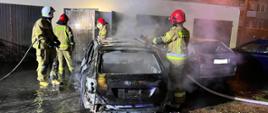 Czterej strażacy gaszą palący się samochód osobowy. Widoczne znaczne zniszczenie pojazdu i unoszący się dym.