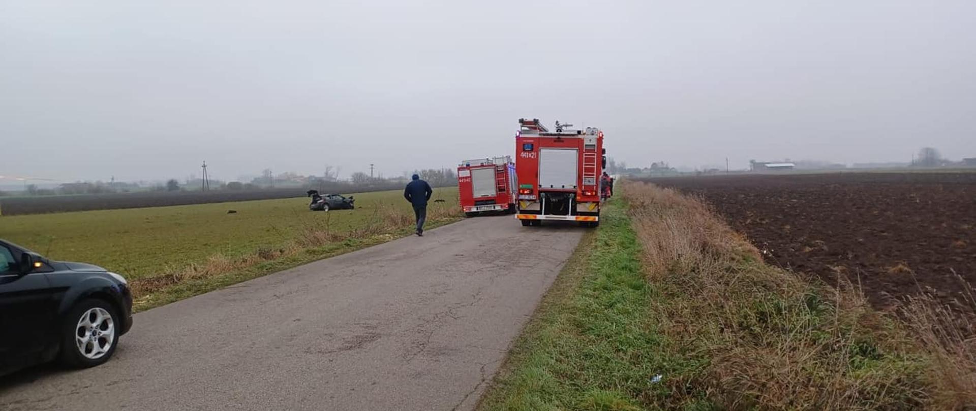 Na zdjęciu widać dwa czerwone samochody strażackie stojące na asfaltowej drodze. Po lewej stronie widać część samochodu osobowego. Drogą w stronę samochodów strażackich idzie mężczyzna ubrany w ciemny strój. Na głowie ma zimową czapkę. Obok samochodu strażackiego stoi ratownik medyczny. W oddali widać rozbity samochód osobowy, który stoi na polu. W tle widać zabudowania oraz pola. Jest widno.